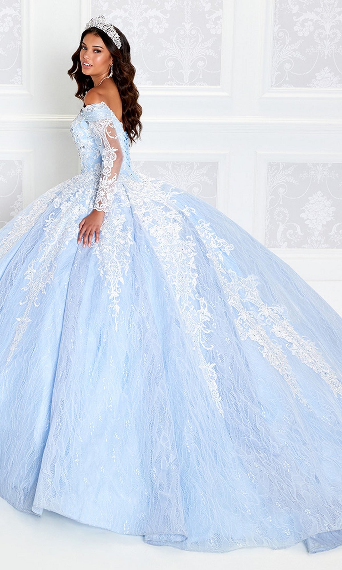 A woman wearing the Princesa Off-the-Shoulder Lace Quince Dress PR12267 with a lace-up corset back and tiara stands in an elegant, white-paneled room.